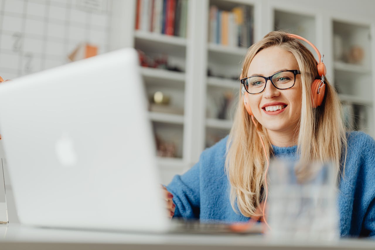Young woman with glasses and headphones smiling while using a laptop indoors.